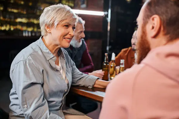Feliz mulher de meia-idade ouvindo colega barbudo perto turvo equipe multiétnica falando no pub — Fotografia de Stock