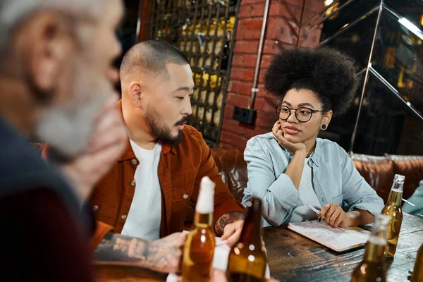 Mujer afroamericana escuchando a colegas multiétnicos discutir nuevo proyecto de negocio en el pub - foto de stock