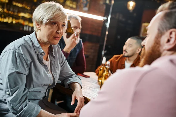 Smiling middle aged woman listening to colleague near blurred multiethnic workmates talking in pub — Stock Photo