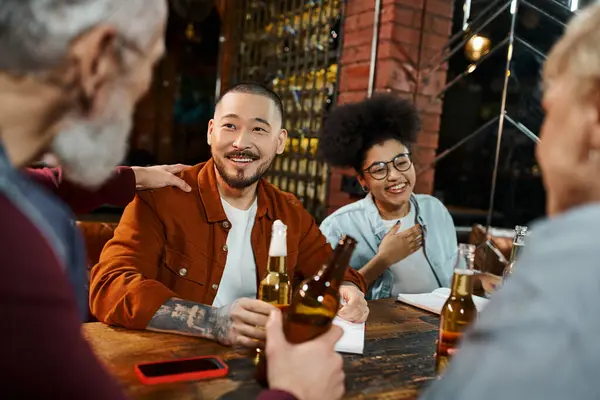 Alegres colegas de negocios multiétnicos sosteniendo botellas de cerveza y hablando de un nuevo proyecto en el pub - foto de stock