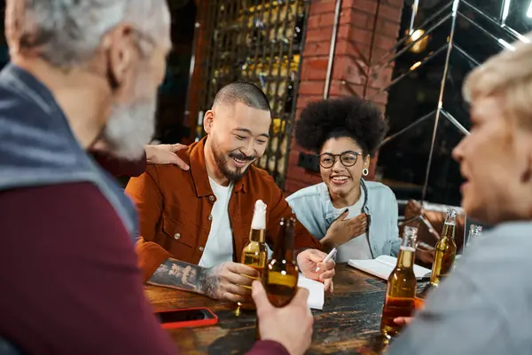 Радісні багатоетнічні бізнесмени тримають пляшки пива і говорять про новий бізнес-проект у пабі — стокове фото