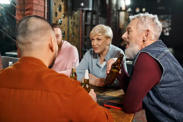 Bearded businessman holding beer bottle and talking to multicultural team resting in pub after work — Stock Photo