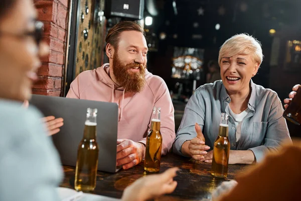 Hombre barbudo y mujer de mediana edad riendo cerca del portátil y la mujer afroamericana hablando en el pub - foto de stock