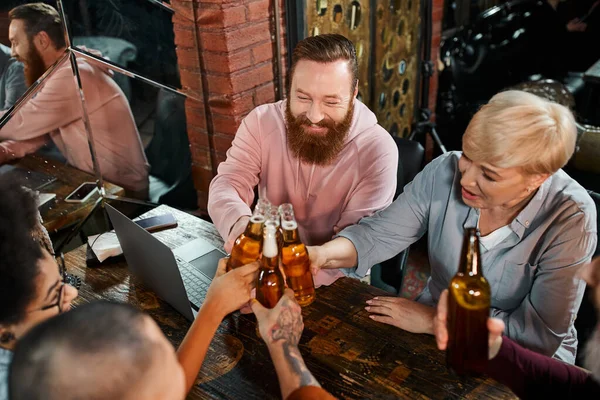 Successful business colleagues clinking beer bottles while sitting near laptop in pub after work — Stock Photo