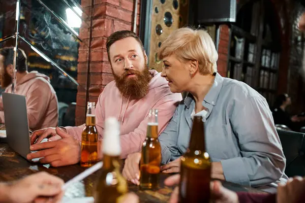 Mujer de mediana edad mirando portátil cerca inspirado colega mientras está sentado cerca de botellas de cerveza en el pub - foto de stock