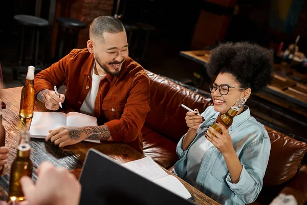 Cheerful african american woman with beer bottle near asian colleague sitting with notebook in pub — Stock Photo