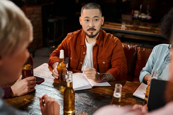 Tatuado asiático hombre sentado con cerveza y notebook cerca multicultural compañeros de trabajo en pub después de trabajo - foto de stock