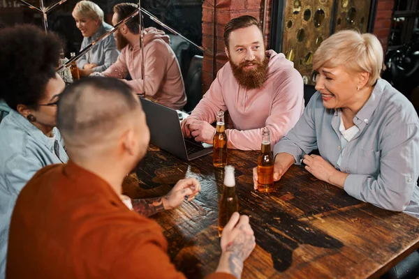 Femme d'âge moyen riant près de collègues multiethniques à côté de l'ordinateur portable et des bouteilles de bière dans le pub — Photo de stock