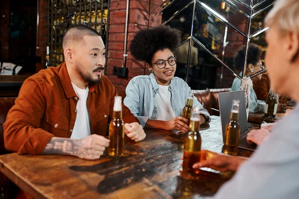 Feliz equipo diverso de compañeros de trabajo multiétnicos discutiendo puesta en marcha cerca de botellas de cerveza y portátil en el pub - foto de stock