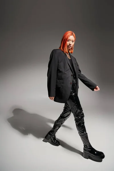 Стильная и уверенная в себе азиатская женщина с рыжими волосами, ходящими в черном одеянии, в латексных брюках и блейзере — стоковое фото