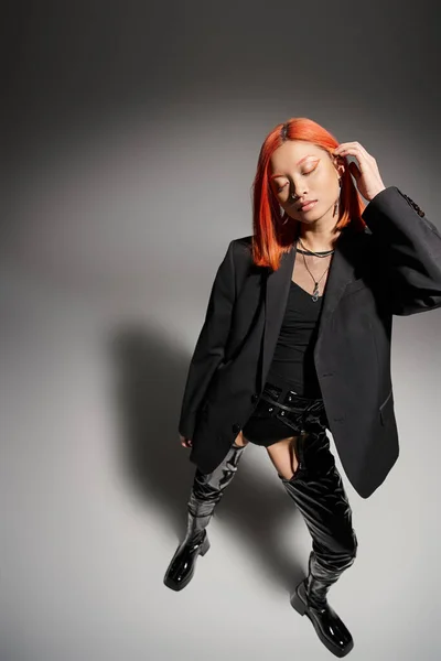 Stylish asian woman with red hair posing in black latex boots and blazer on grey background — Stock Photo