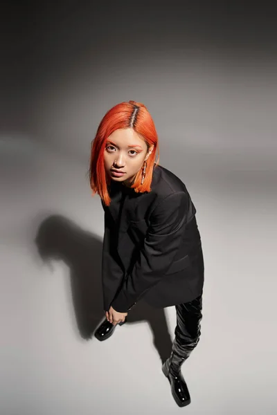 Asian woman with red hair posing in black latex boots and blazer on grey background, high angle — Stock Photo