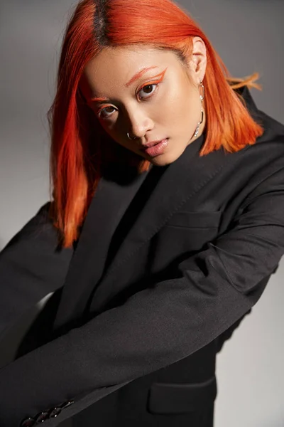 Asiatin mit roten Haaren und orangefarbenem Eyeliner posiert im schwarzen Blazer auf grauem Hintergrund, Stil — Stockfoto