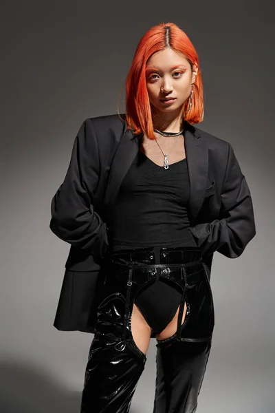 Сором'язлива молода азіатська жінка в сексуальному одязі з латексними черевиками і блейзер позує на сірому тлі, мода — стокове фото