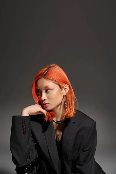 Fashionable asian woman with piercing in nose and red hair posing in blazer on grey background — Stock Photo