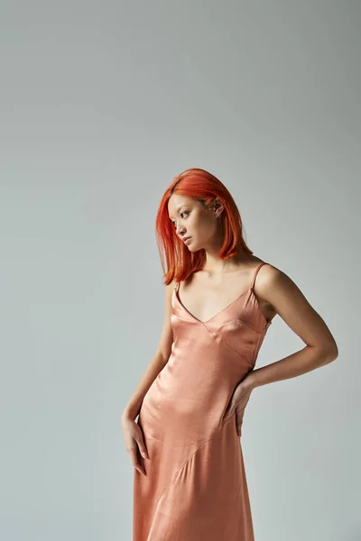 Sensual young asian woman with red hair posing in slip dress with hand on hip on grey backdrop — Stock Photo