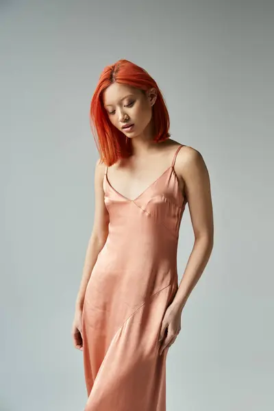 Sensual young asian woman with red hair posing in pastel slip dress on grey backdrop, looking away — Stock Photo