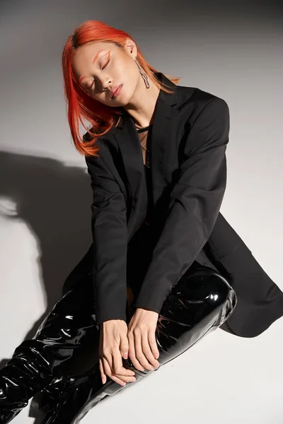 Fashionable asian woman in sexy latex boots and blazer sitting with closed eyes on grey backdrop — Stock Photo