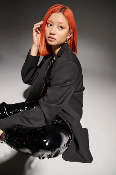 Молодая азиатская женщина с пирсингом носа регулируя рыжие волосы и сидя в дерзких латексных сапогах на сером — стоковое фото