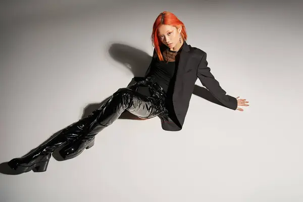 Mode edgy, femme asiatique aux cheveux roux posant en tenue audacieuse sur fond gris, bottes en latex noir — Photo de stock