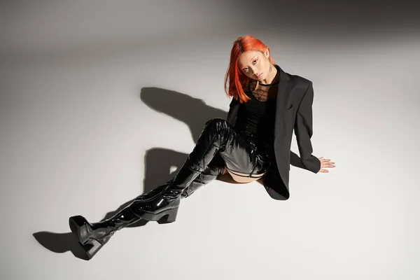 Edgy fashion, asian woman with red hair sitting in bold outfit on grey background, black latex boots — Stock Photo