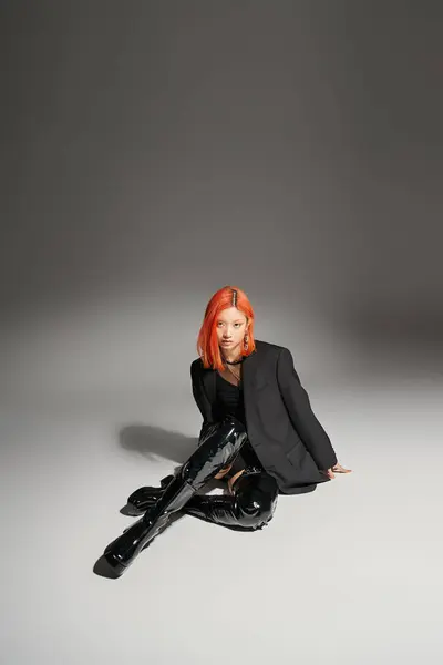 Modelo asiático seguro de sí mismo con el pelo rojo sentado en botas de látex negro y chaqueta sobre fondo gris - foto de stock