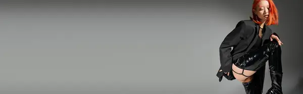 Giovane modella asiatica con capelli rossi in posa in stivali di lattice e blazer oversize su sfondo grigio, banner — Foto stock