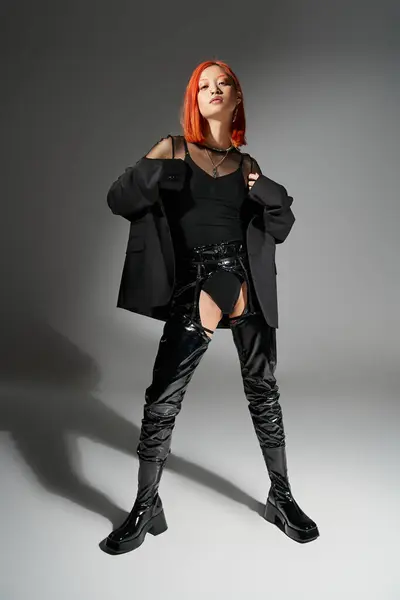 Стильная азиатская женщина с рыжими волосами позирует в блейзере и черных ботинках из латекса на сером фоне — стоковое фото