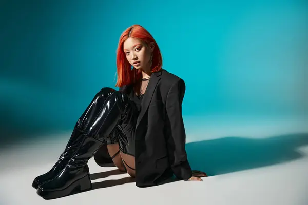 Asian model with nose piercing sitting in oversized blazer and black latex boots on blue backdrop — Stock Photo