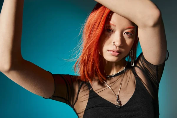 Mujer asiática con piercing en la nariz posando con las manos levantadas y blusa transparente sobre fondo azul - foto de stock