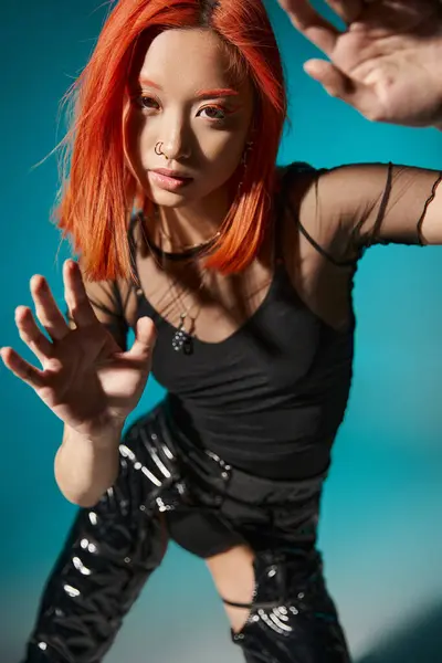 Asian model with piercing and red dyed hair gesturing and looking at camera on blue background — Stock Photo