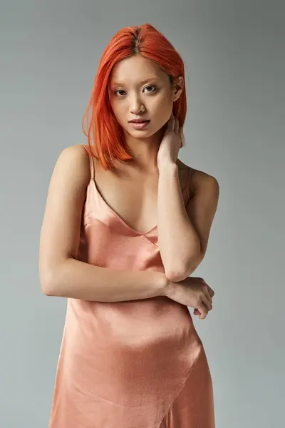Hermosa y joven asiático mujer en pastel rosa slip vestido posando sobre gris fondo, mira la cámara - foto de stock