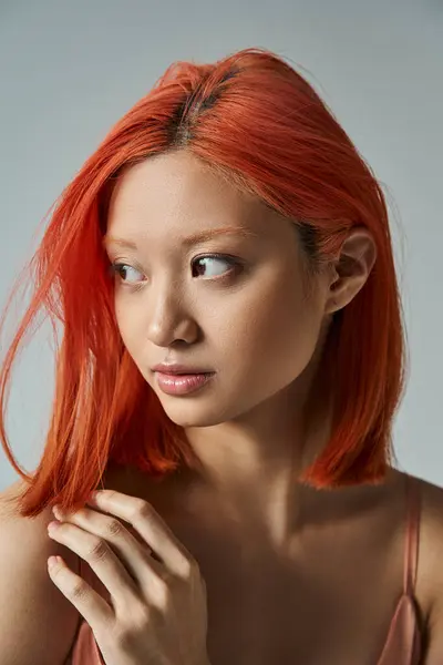 Elegante y joven mujer asiática con el pelo rojo y maquillaje natural mirando hacia otro lado en el fondo gris - foto de stock