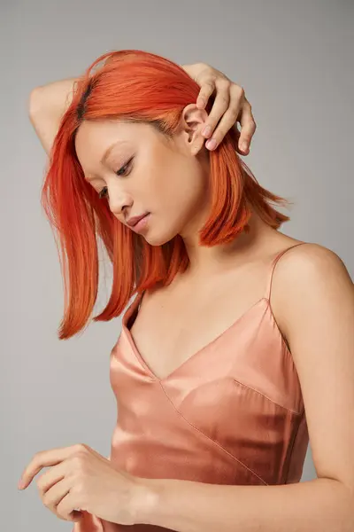 Портрет молодой азиатской женщины с идеальной кожей, регулирующей рыжие волосы на сером фоне, женская грация — стоковое фото