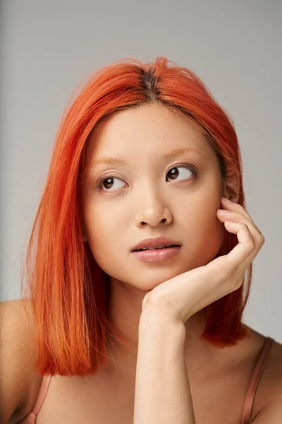 Retrato de mujer asiática atractiva y joven con la piel perfecta soñando despierto sobre fondo gris - foto de stock