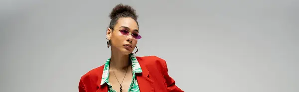 Mujer afroamericana con estilo en gafas de sol de color rosa y ropa vibrante de moda posando en gris, bandera - foto de stock