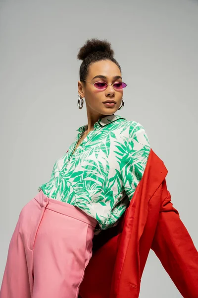 Африканская американка в солнцезащитных очках и модной одежде позирует на сером фоне, в индивидуальном стиле — стоковое фото