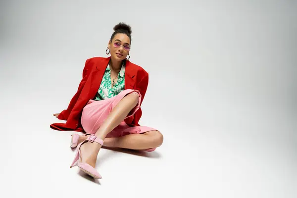 Модна афроамериканка в яскравому одязі і рожевих сонцезахисних окулярах, що сидять на сірому тлі — стокове фото