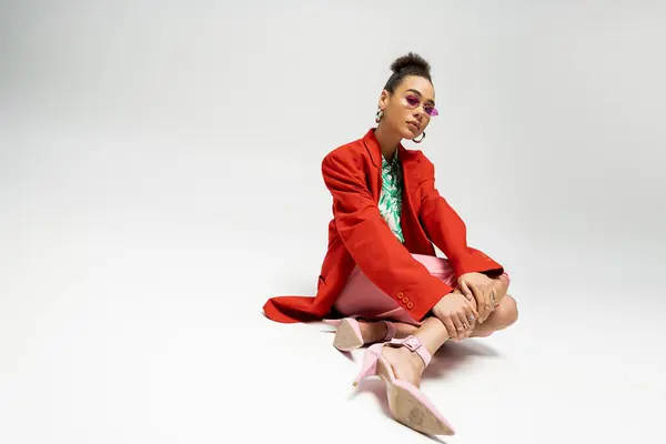 Mujer afroamericana en traje vibrante y gafas de sol rosadas sentadas sobre fondo gris, trendsetter - foto de stock