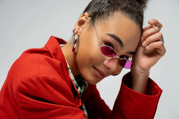 Portrait du modèle afro-américain positif en tenue élégante et lunettes de soleil roses sur fond gris — Photo de stock