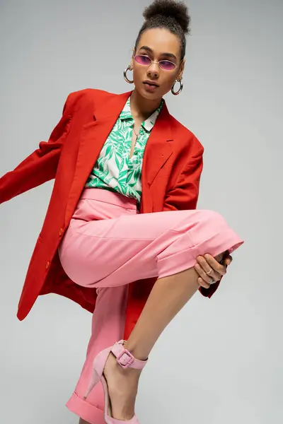Modisches afrikanisch-amerikanisches Model in fetter Kleidung und Sonnenbrille vor grauem Hintergrund, dynamische Pose — Stockfoto