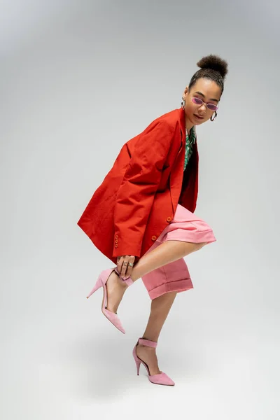 African american fashion model in red blazer adjusting strap on high heels on grey backdrop — Stock Photo