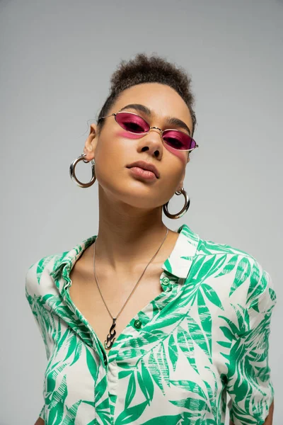 African american woman in pink sunglasses and hoop earrings looking at camera on grey backdrop — Stock Photo