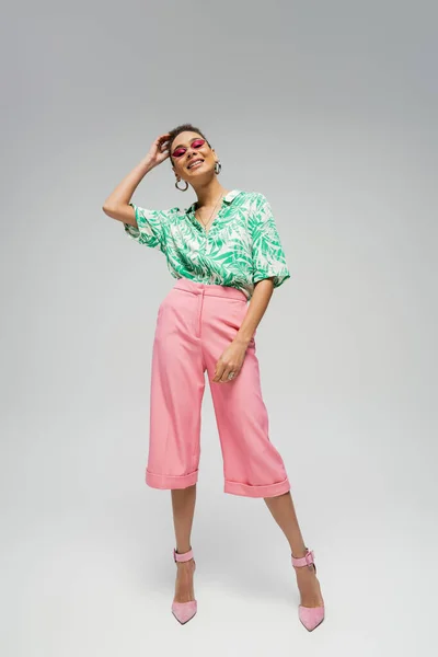 Счастливая африканская американка в розовых солнцезащитных очках и стильной одежде позирует на сером фоне — стоковое фото