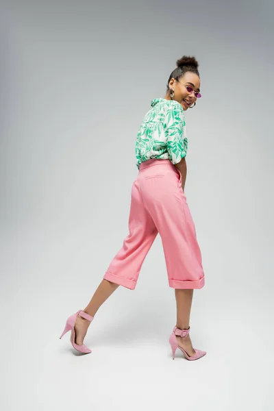 Щаслива афроамериканка в рожевих сонцезахисних окулярах і стильний одяг, що йде на сірому фоні — стокове фото