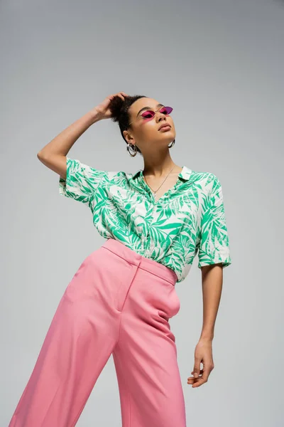 Joven modelo afroamericano de moda en traje de moda y gafas de sol de color rosa posando sobre fondo gris - foto de stock