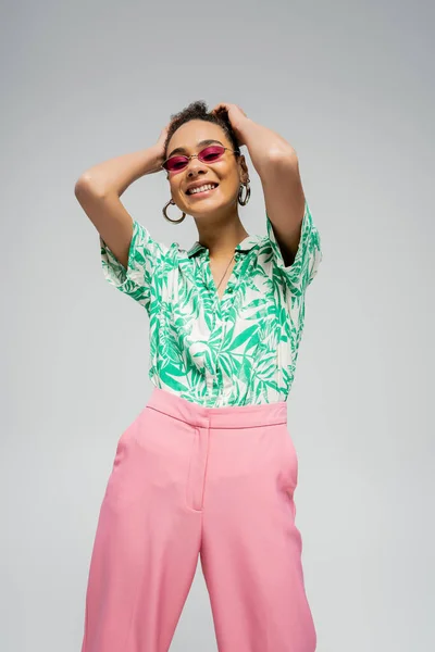 Jovem modelo de moda afro-americana em trajes da moda e óculos de sol rosa sorrindo em pano de fundo cinza — Fotografia de Stock