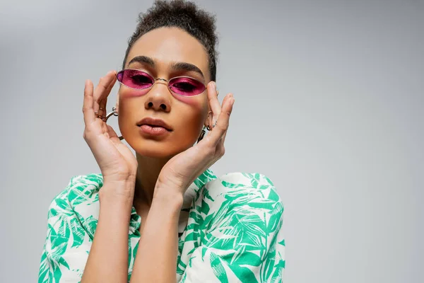 Кучерява афроамериканка в рожевих сонцезахисних окулярах і позує в стильному вбранні на сірому фоні — стокове фото