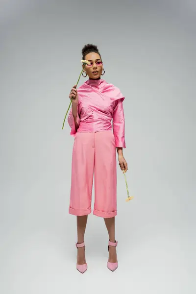 Повна довжина стильної афроамериканської жінки в рожевому одязі і сонцезахисних окулярах позує з квітами — стокове фото