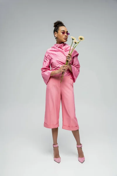 Piena lunghezza di donna afroamericana in abito rosa e tacchi in posa con fiori su sfondo grigio — Foto stock